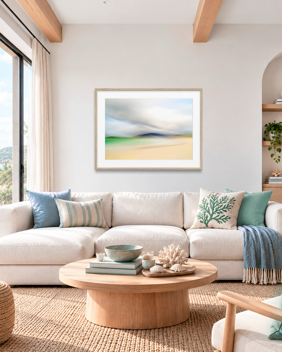 A framed abstract landscape art print hangs on a white wall above a cream sectional sofa in a bright, modern living room. The print features soft, blended colors of blue, green, and beige, styled with neutral-toned pillows and a round wooden coffee table in the foreground.