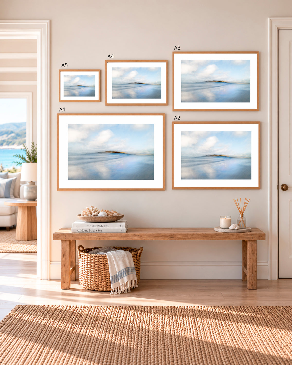 Five framed abstract landscape prints in varying sizes are arranged on a beige wall above a wooden bench in a styled room. The prints feature soft blue, white, and gray water-like imagery, and the room has a woven rug, basket, and coastal decor visible through an open doorway.