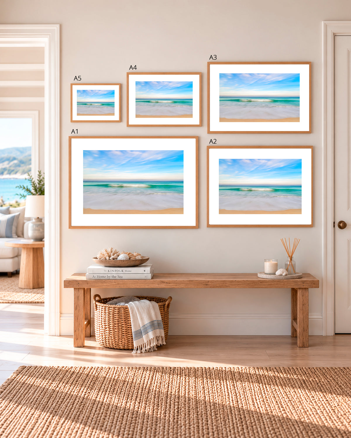 A staged interior room showing a gallery wall with five framed beach prints in different sizes, mounted above a wooden bench with decorative items. The prints depict blue sky, white waves, and pale sand, and the room has a warm neutral wall and rug.