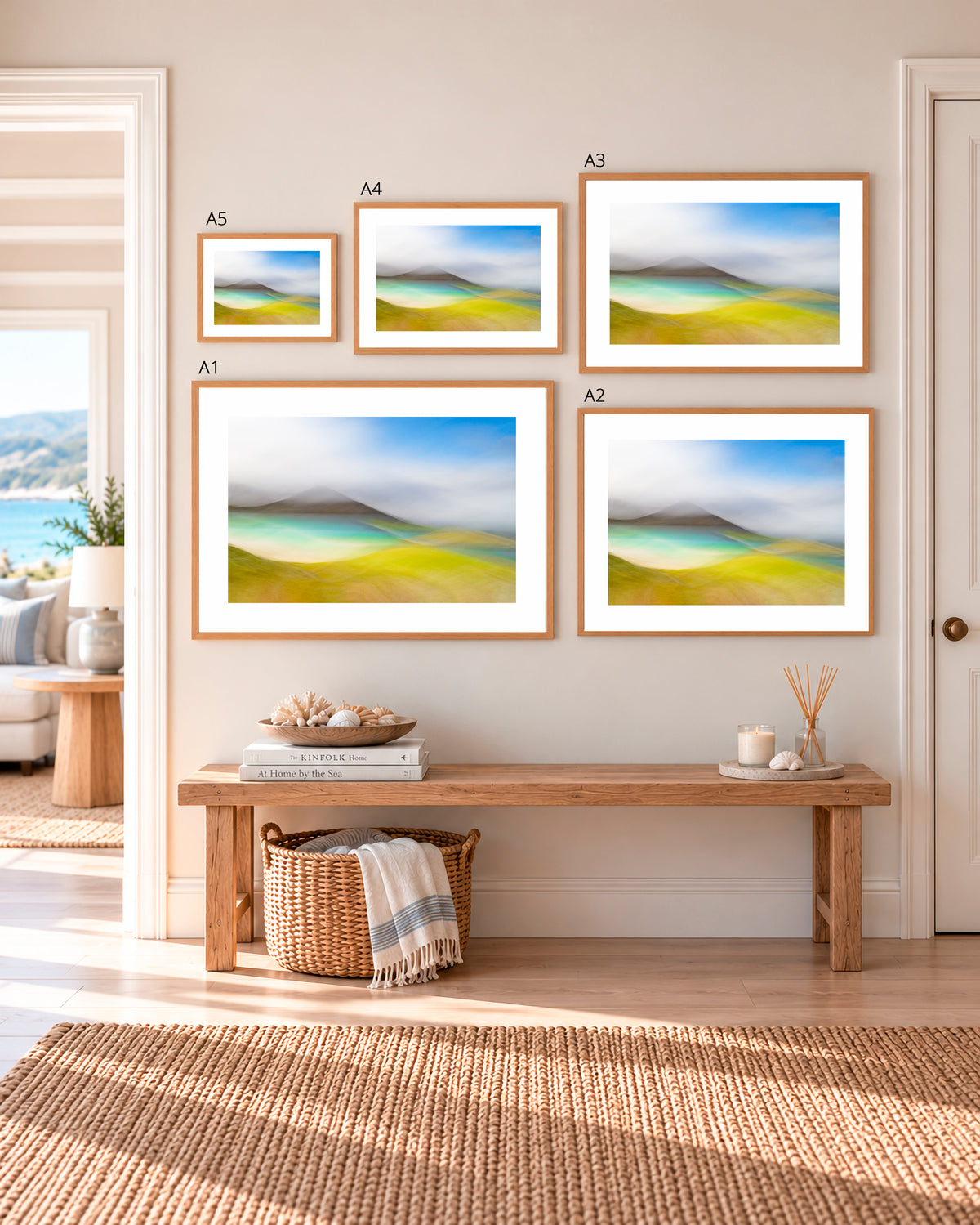 Five framed landscape art prints in different sizes are displayed on a beige wall above a wooden bench in a bright, modern room. The prints depict soft, hazy coastal hills with green fields and blue skies.