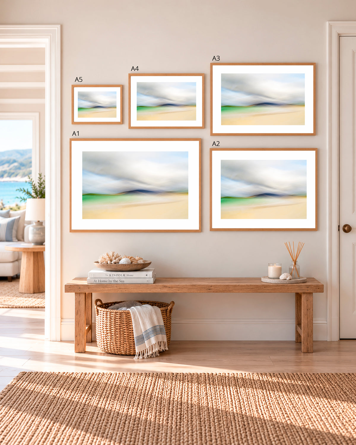Five framed abstract beach landscape prints in varying sizes are arranged on a beige wall above a wooden bench in a bright, modern room. The prints feature soft, blended tones of blue, green, beige, and white, and are presented with white mats and light wood frames.