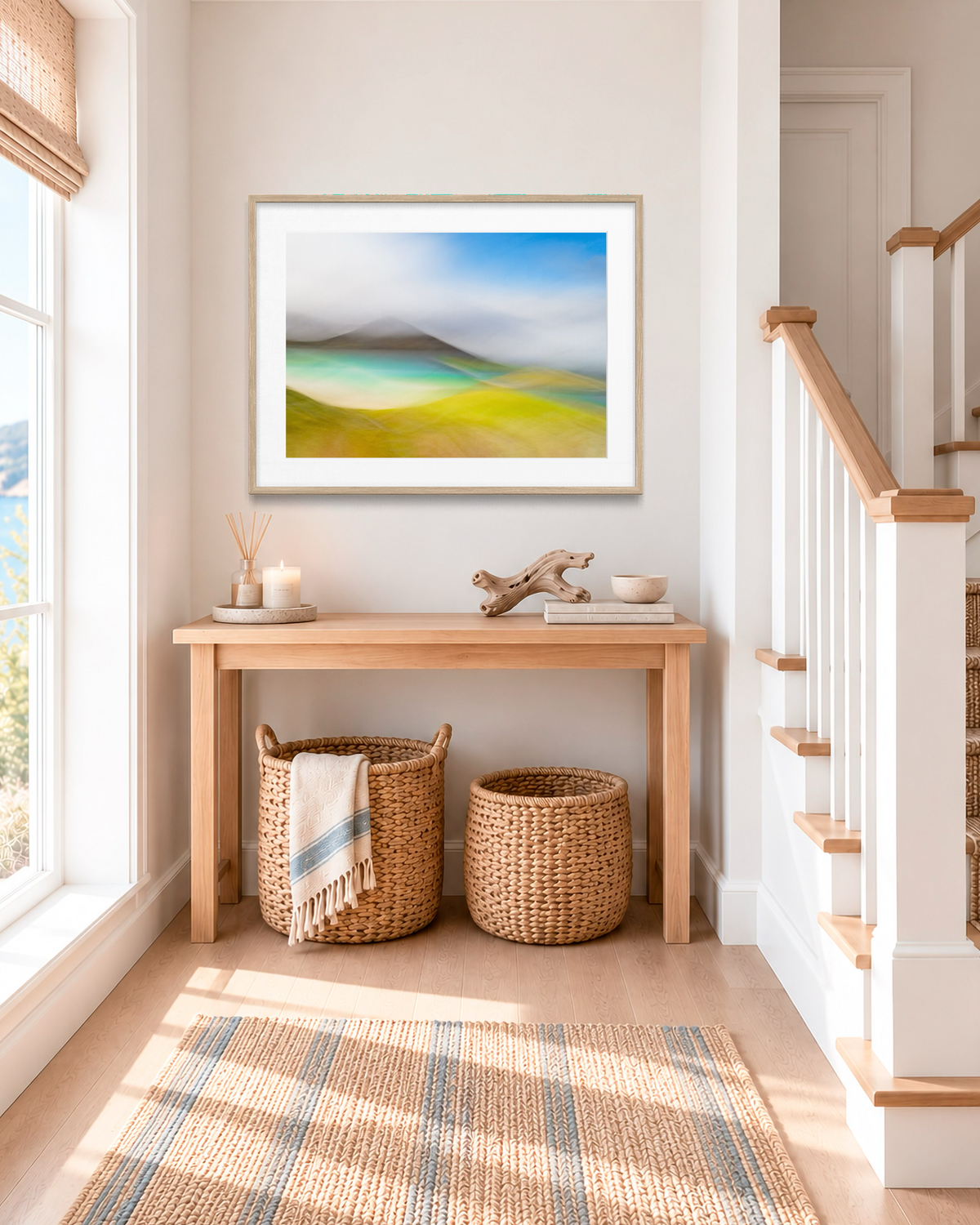 A framed abstract landscape art print hangs on a light beige wall above a wooden console table in a bright, modern entryway. The print features soft gradients of blue, green, and yellow.