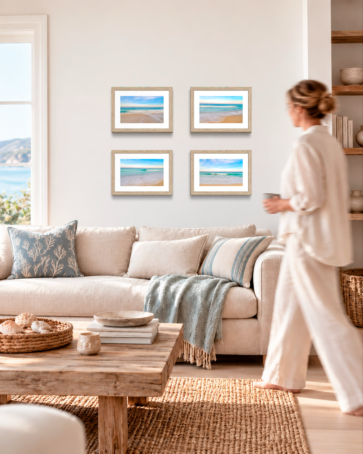 A woman in a cream lounge set walks past a beige sofa in a bright, modern living room. The wall above the sofa features a 2x2 grid of four framed beach prints with light wood frames.