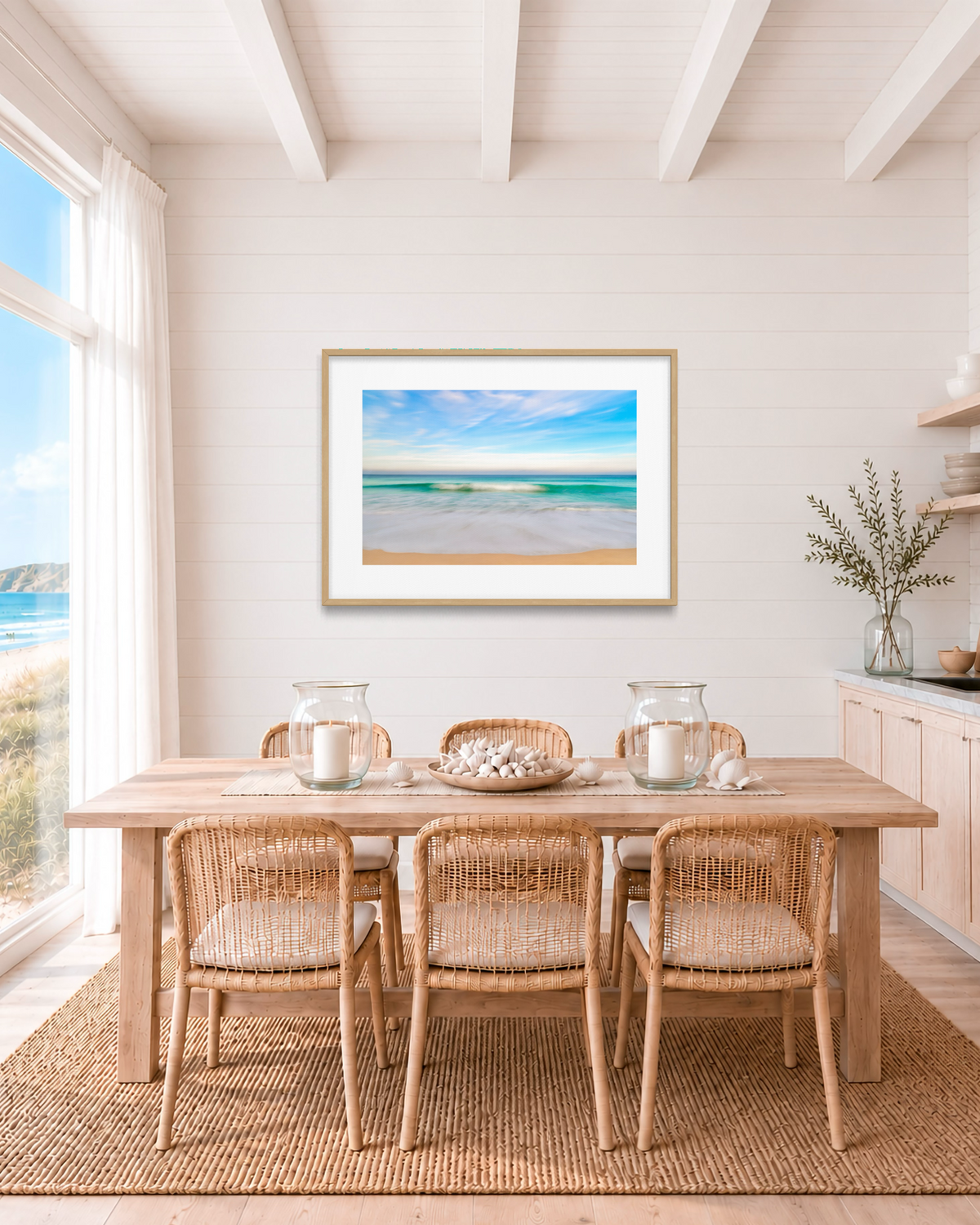 A framed beach landscape print hangs on a white shiplap wall above a wooden dining table and woven chairs in a bright, coastal-style dining room.